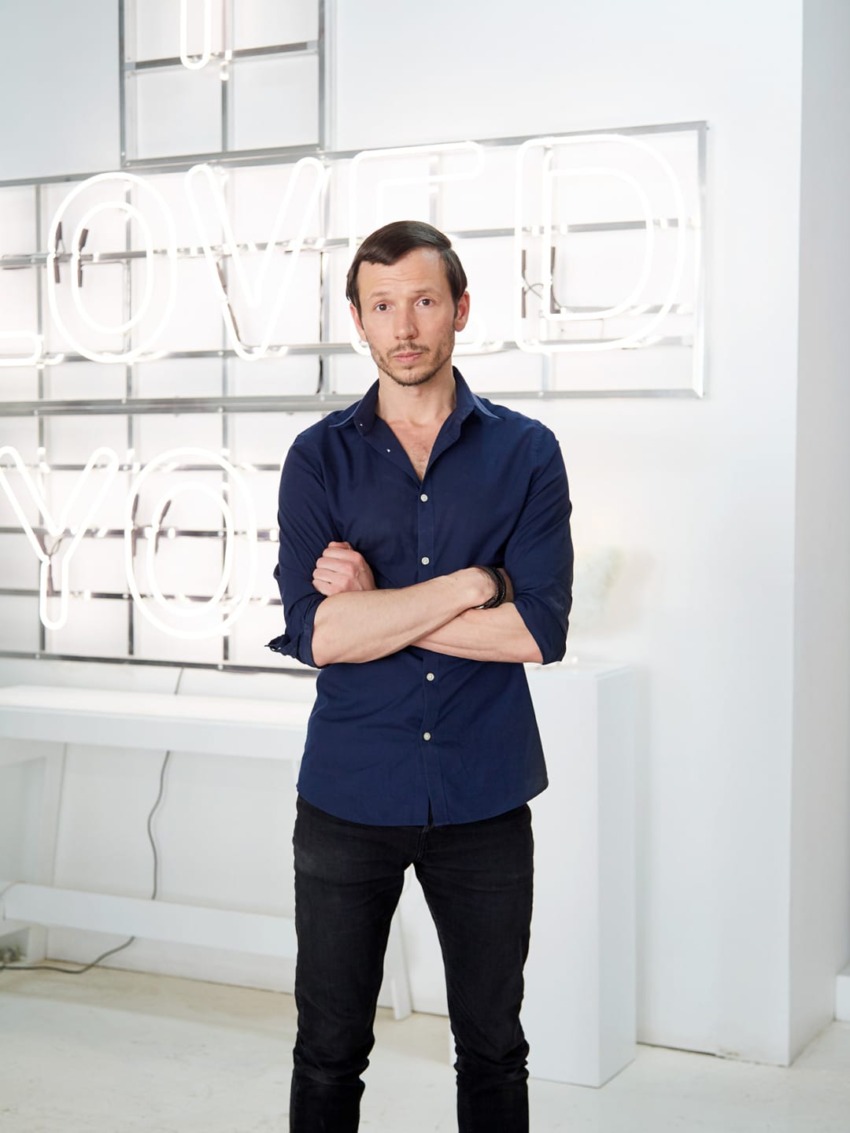 Person in a navy shirt and black pants standing with arms folded in front of a neon "I LOVED YOU" sign.