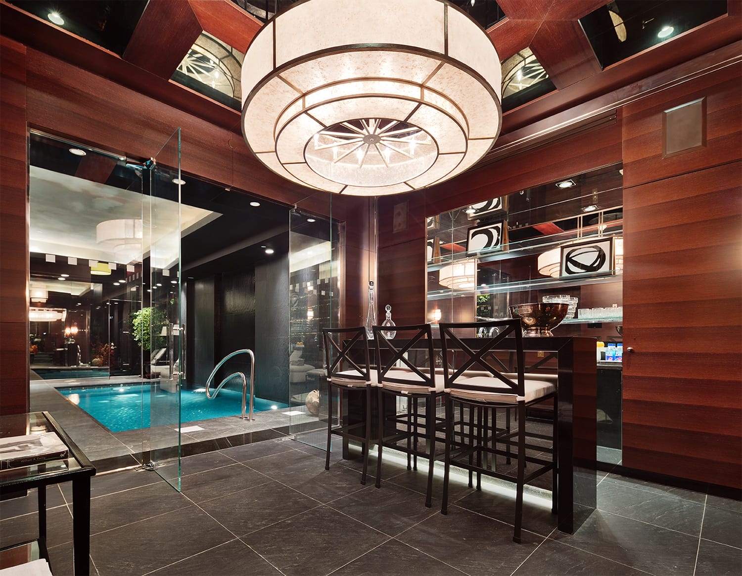 Luxurious indoor swimming pool area with modern wooden decor, seating, large ceiling light, and glass walls.