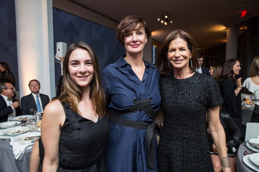 Three people posing together at a formal event, with a dinner table and guests in the background.