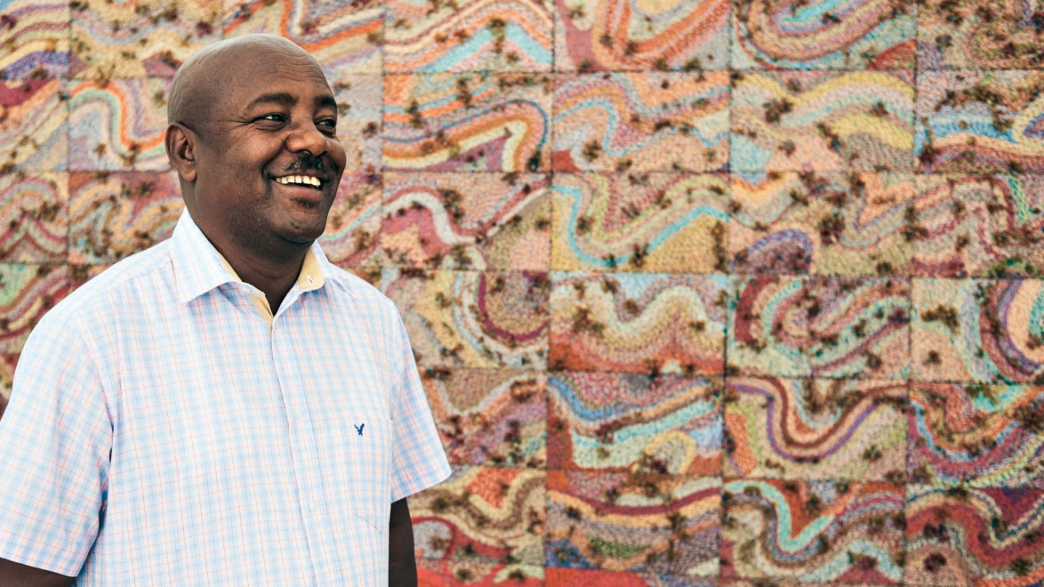 Man smiling in front of a vibrant, colorful abstract mural with a checkered shirt.