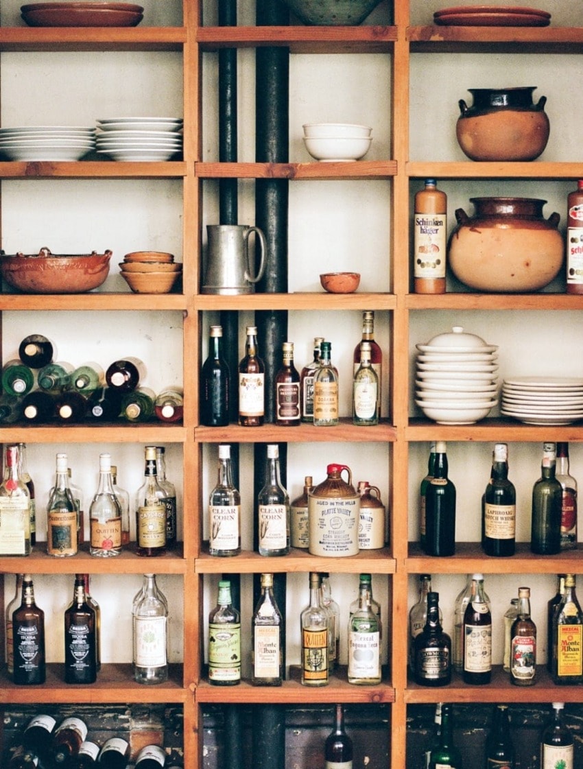 Wooden shelves with assorted bottles, stacked plates, and bowls in a rustic kitchen setting.