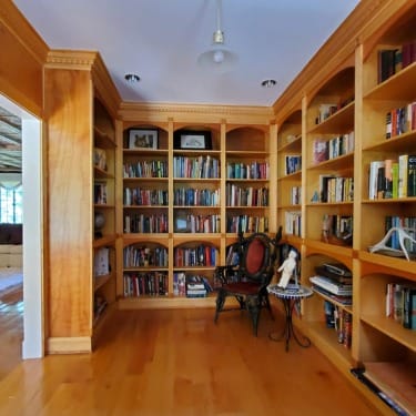 Wooden home library with filled bookshelves, a vintage chair, and a small table with a sculpture, located in a corner room.
