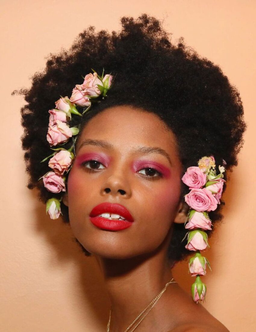 Person with natural hairstyle adorned with pink roses, wearing vibrant makeup against a peach background.