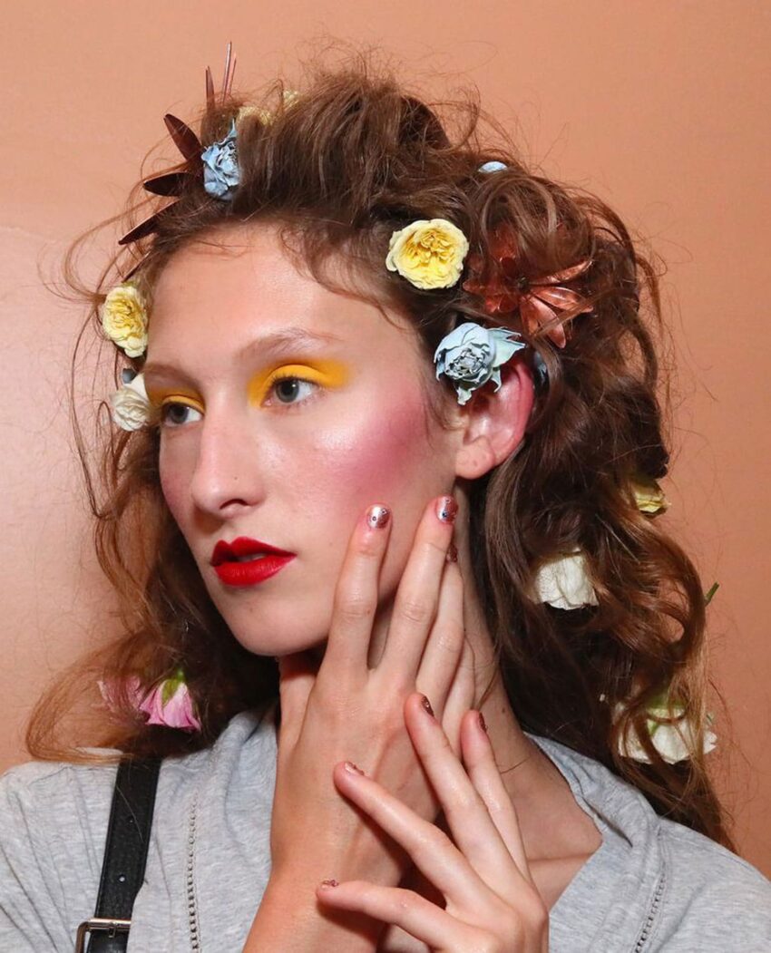 Model with colorful eyeshadow, red lips, and flowers in curly hair touches face against tan background.
