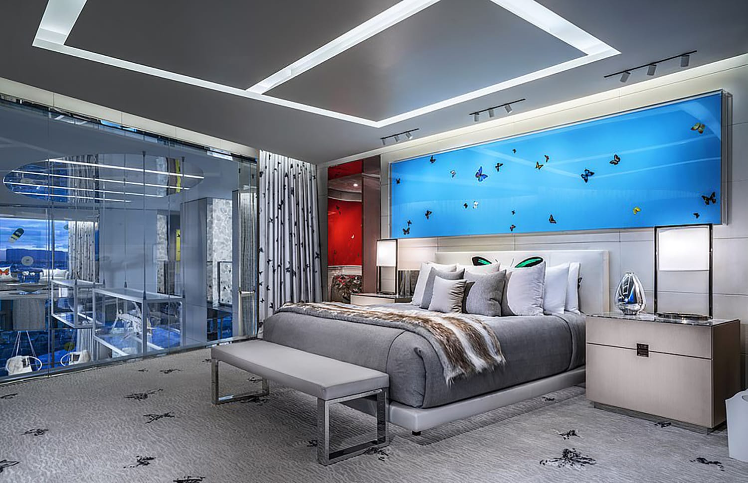 Luxury hotel room with modern decor, stylish gray bed, blue wall art, and glass-walled bathroom in the background.