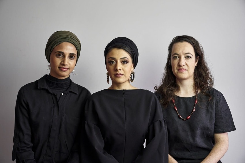Three women wearing black outfits pose against a neutral background.