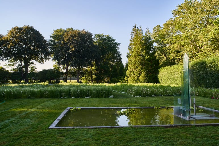 Garden landscape with a reflective pond and modern glass sculpture, surrounded by lush green trees and grass.