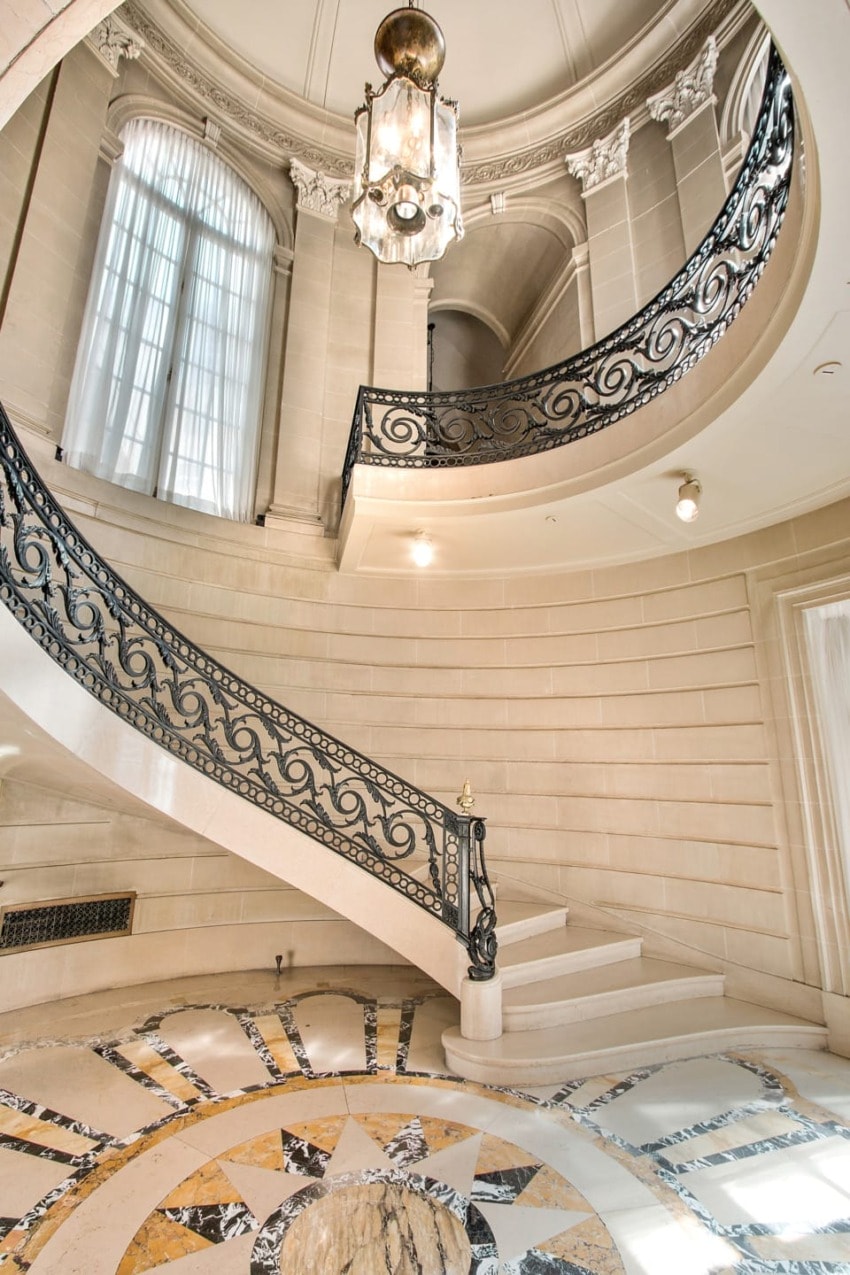 Grand spiral staircase with ornate iron railing, marble floor, and chandelier in elegant interior with large windows.