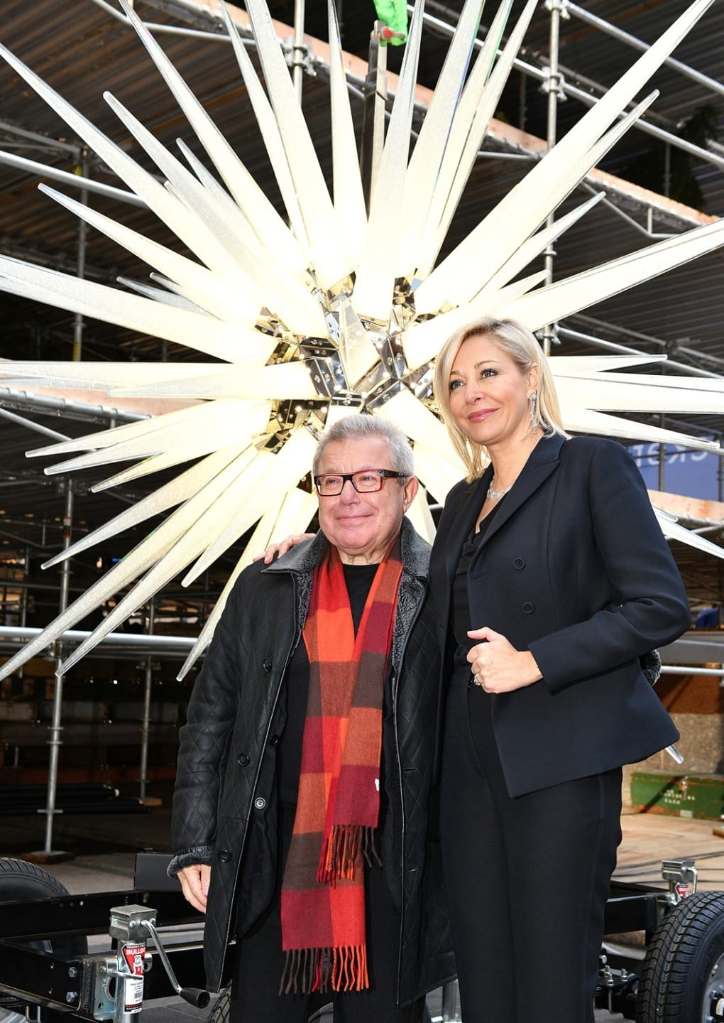 Two people posing in front of a large illuminated starburst installation, wearing winter clothing, with scaffolding in the background.