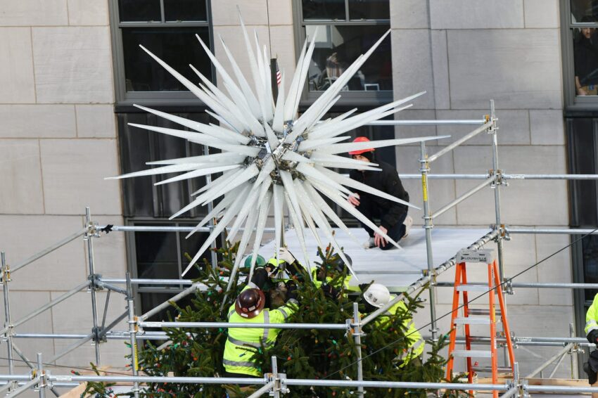 Workers install large star decoration atop Christmas tree using scaffolding.