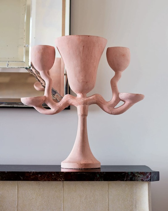 Pink sculptural vase with three branches on a dark marble surface, reflecting in a mirror attached to the wall behind.
