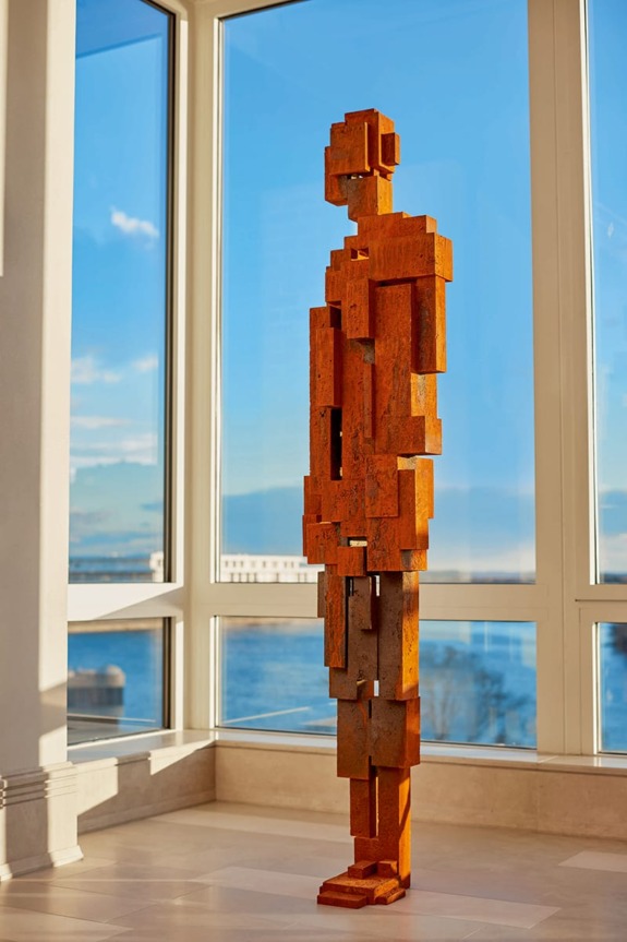Modern abstract metal sculpture in a sunlit room with large windows overlooking a scenic view of water and sky.