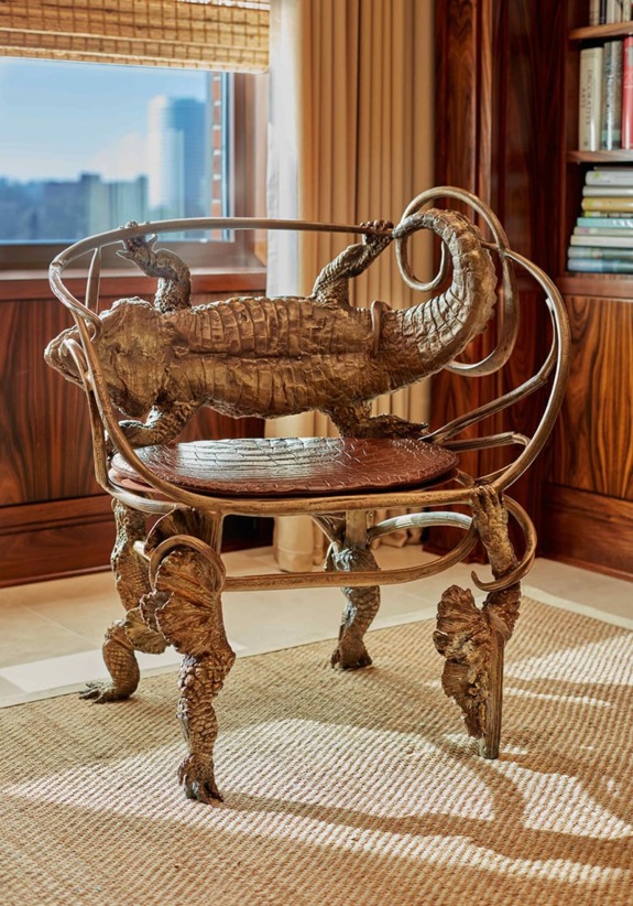 Bronze chair with intricate alligator design, situated in a room with wooden walls and a view of a cityscape outside a window.