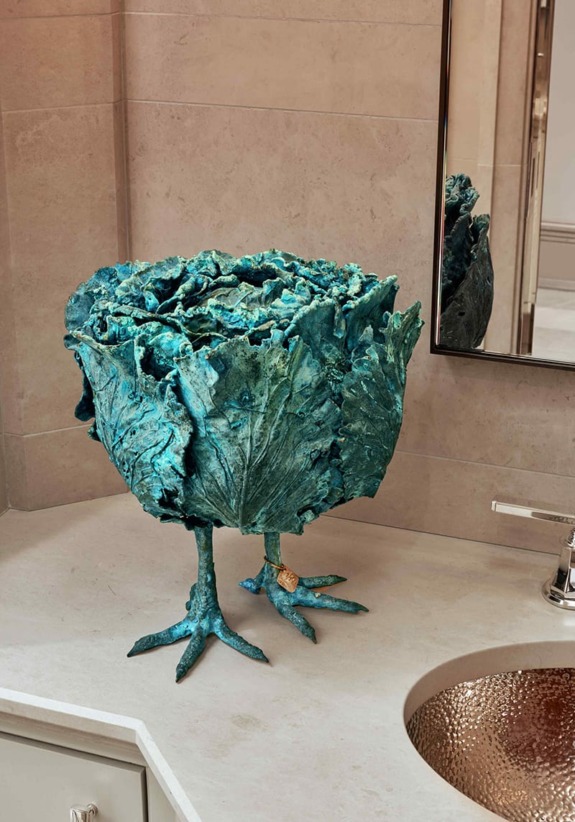 Bronze sculpture of a cabbage with bird legs on a bathroom counter near a copper sink and a mirror.