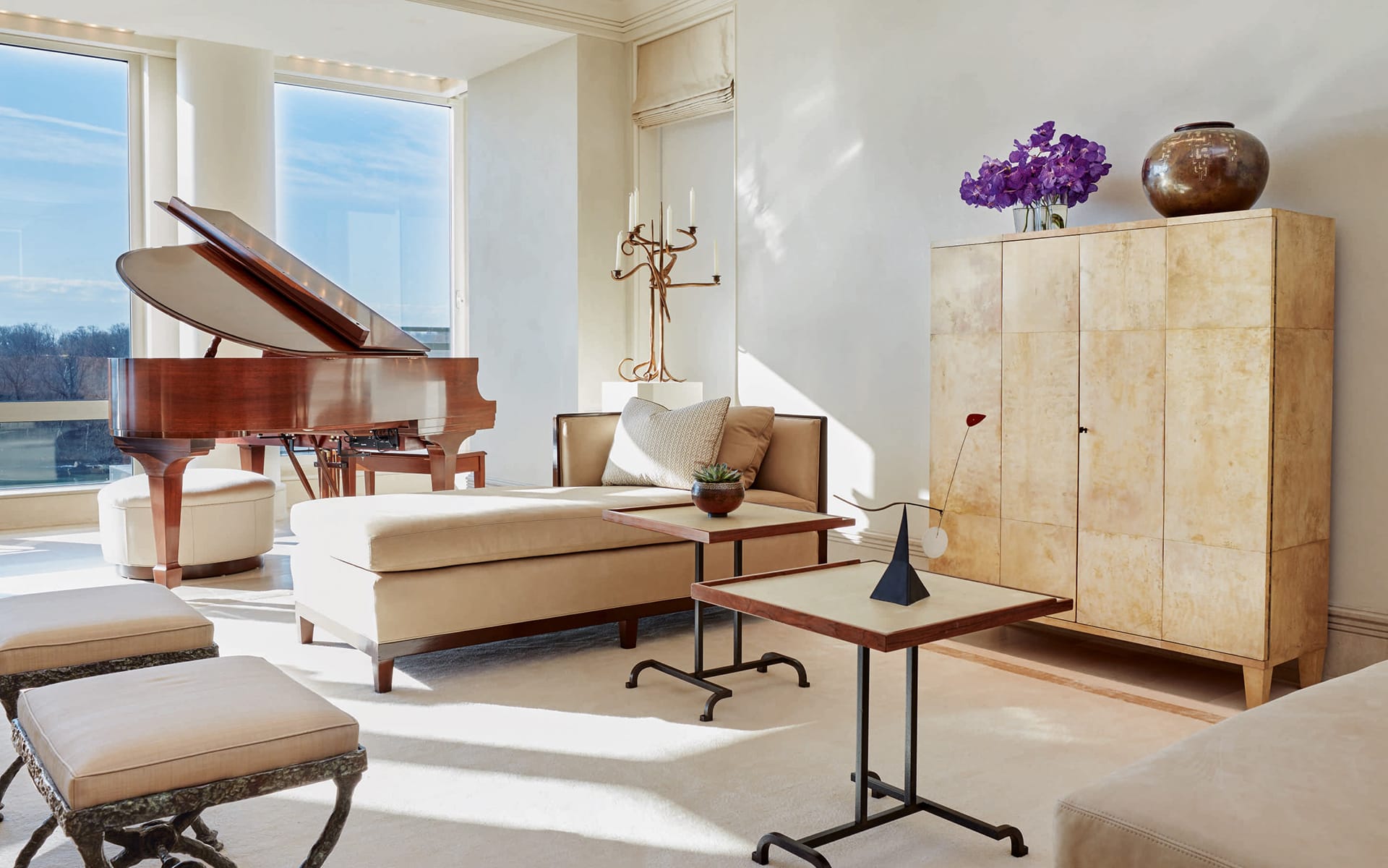 Modern living room with a grand piano, cream furniture, a wooden side cabinet, and large windows letting in natural light.