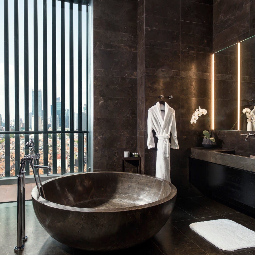 Luxurious bathroom with a large stone tub, a white robe hanging nearby, and a city view through vertical window blinds.
