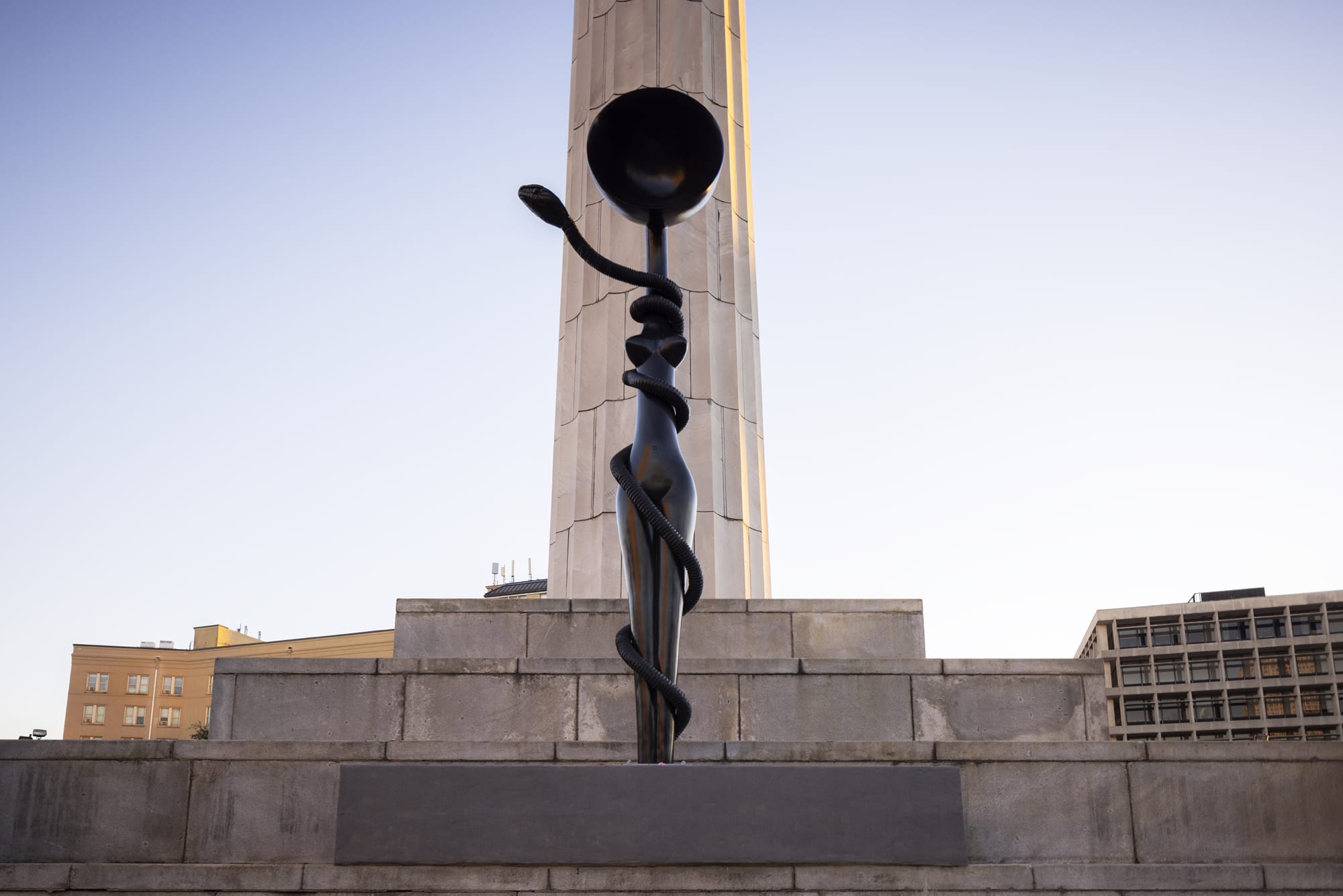 Modern abstract sculpture in city plaza with tall column and blue sky in background