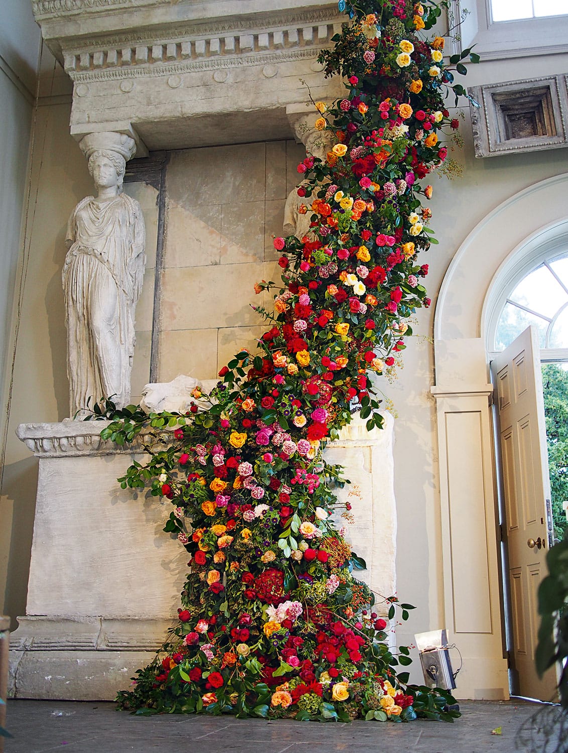 Elegant installation of colorful flowers cascading around a classical statue inside a grand room with large windows.