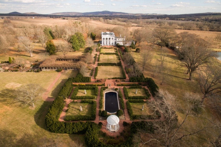 Aerial view of a grand estate with gardens, a reflective pond, and expansive fields in the background.