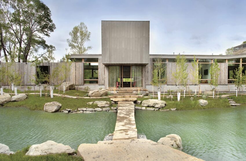 Modern concrete house with a footbridge over a pond, surrounded by trees and a clear sky in the background.