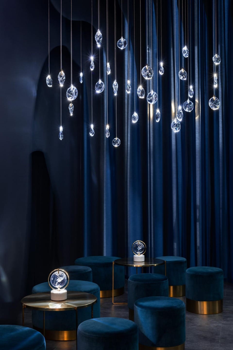 Elegant modern interior with hanging pendant lights, dark blue curtains, and plush teal seating around gold tables.