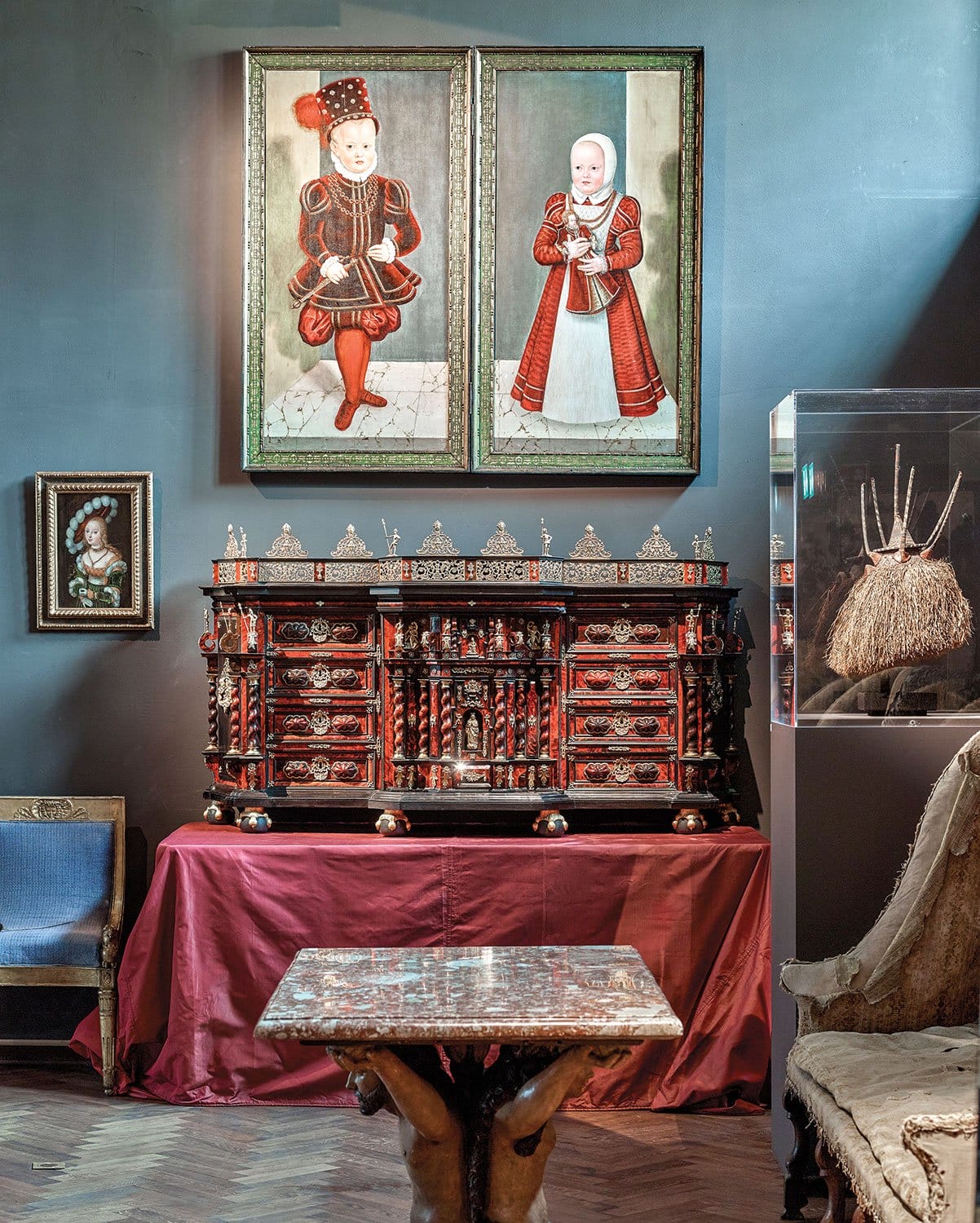 Antique room with ornate cabinet, portraits of children above, and a small table in foreground.