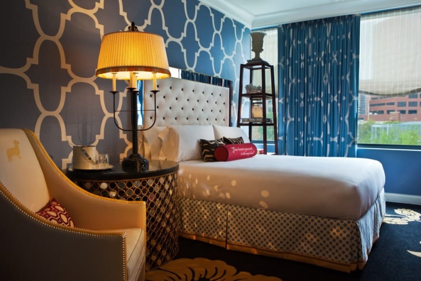Stylish hotel room with patterned blue walls, cozy bed, unique chair, and decorative lamp beside a window with city view.