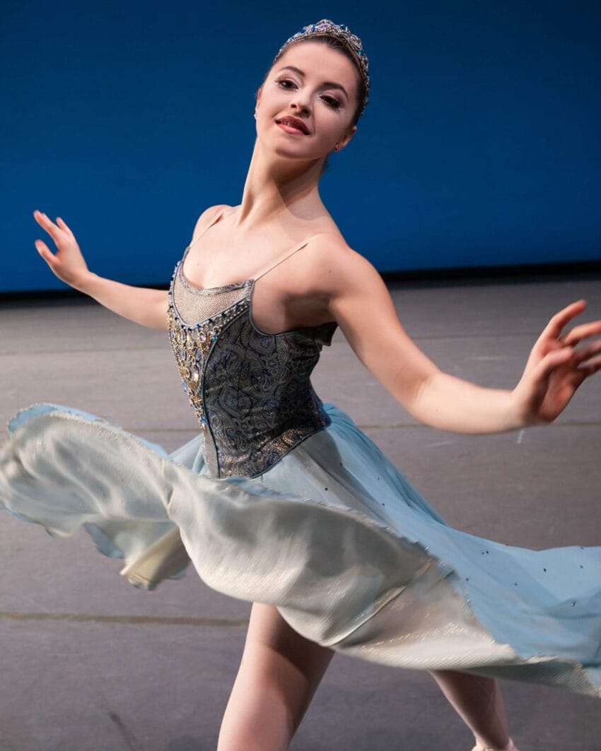 Ballerina gracefully performing a pirouette on stage in a light blue tutu in front of a blue background.