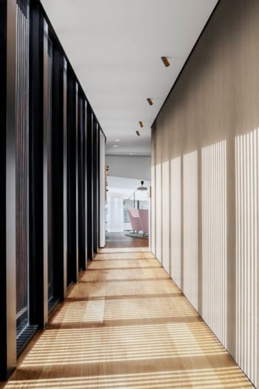 Modern hallway with wooden floor and large vertical windows casting sunlight, leading to a room with pink chairs in the distance.
