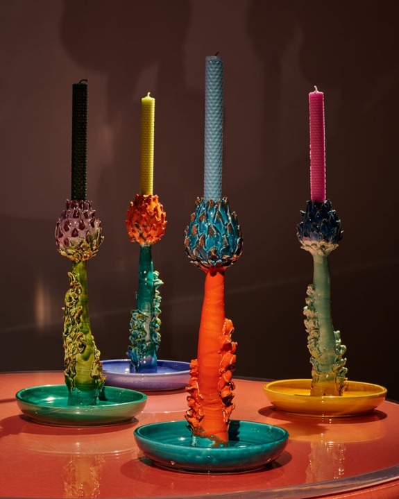 Four colorful, intricately designed candle holders with lit candles, displayed on a reflective pink surface in dim lighting.