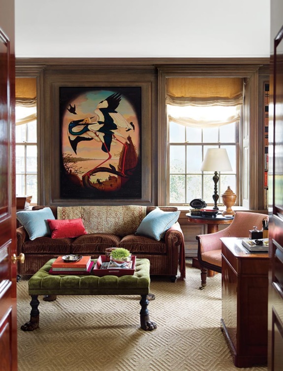 Cozy living room with a painting, sofa, colorful cushions, green footstool, windows, and a wooden desk.