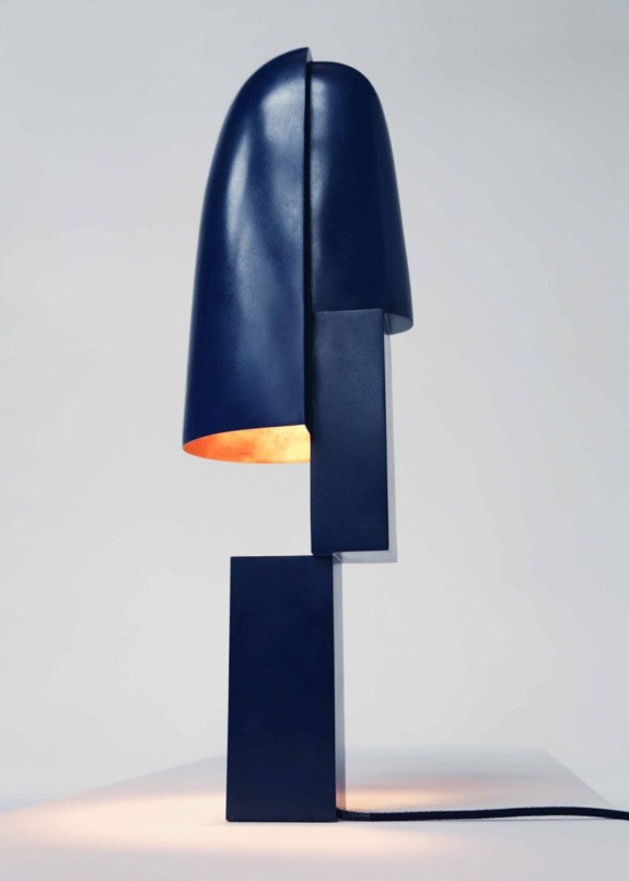 Modern abstract navy blue metal lamp with geometric design and orange interior glow, casting light on a white surface.