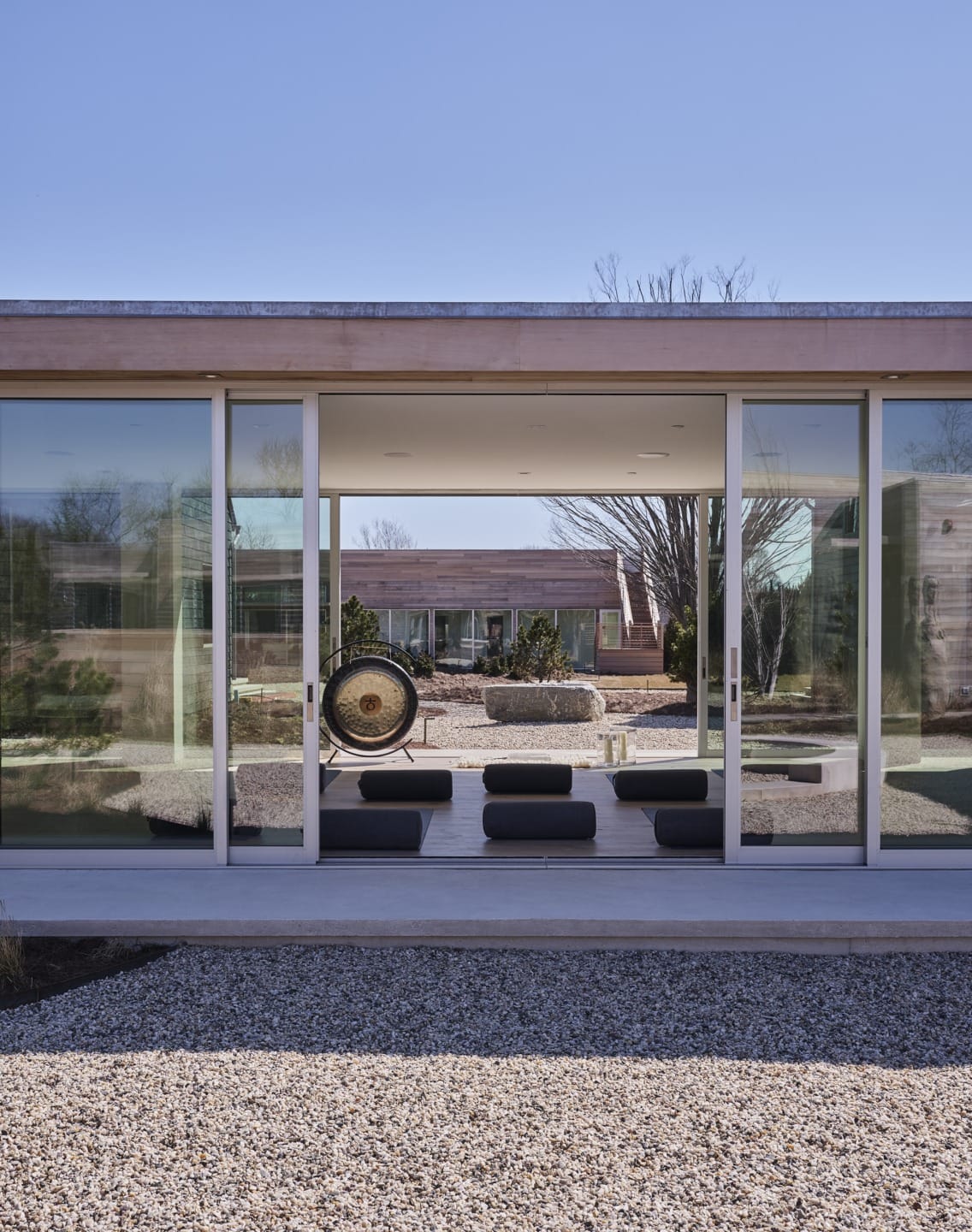 Modern glass house exterior with reflection of trees, minimalist design, outdoor seating, and stone garden in foreground