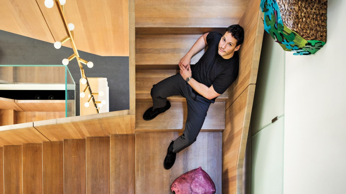 Man sitting on wooden staircase, looking up at camera with modern decor around him and hanging light fixtures visible.