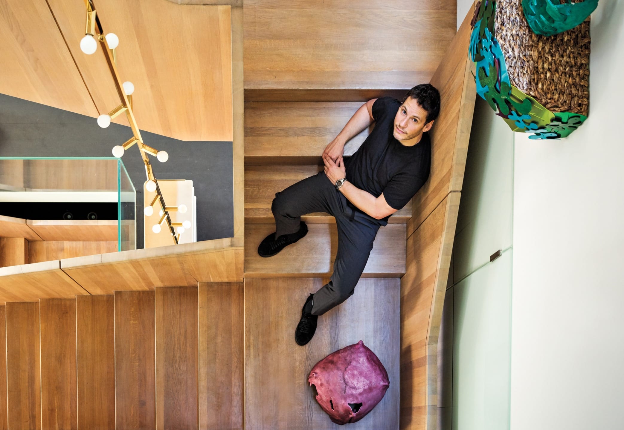 Man sitting on wooden staircase, looking up at camera with modern decor around him and hanging light fixtures visible.