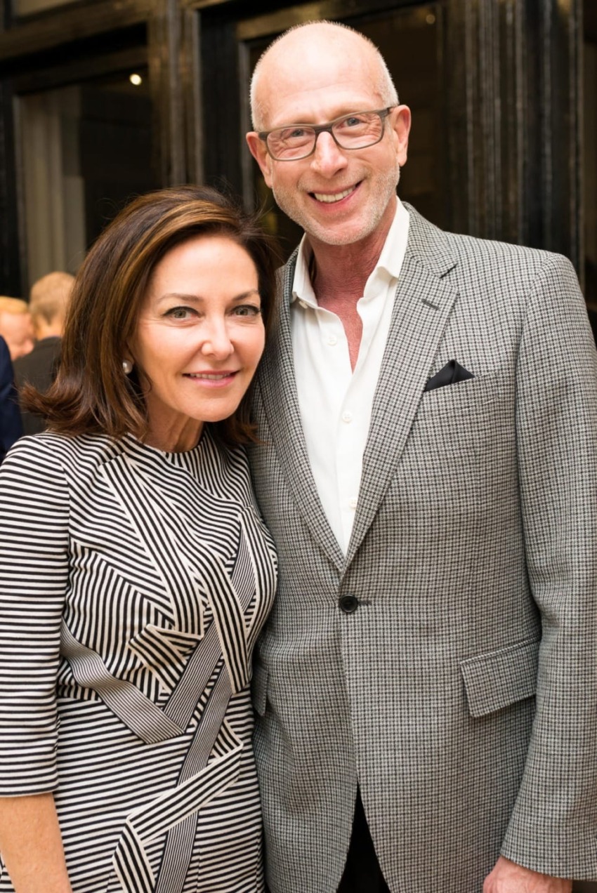 A smiling couple poses together at an indoor event, both wearing fashionable clothing.