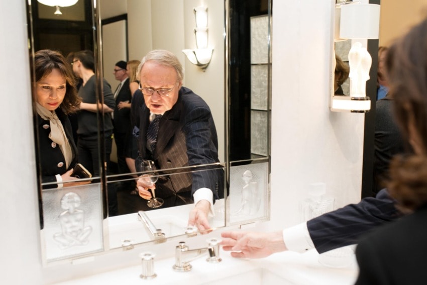 Man in suit holding wine glass, reaching towards bathroom sink, with woman watching and group of people in the background.