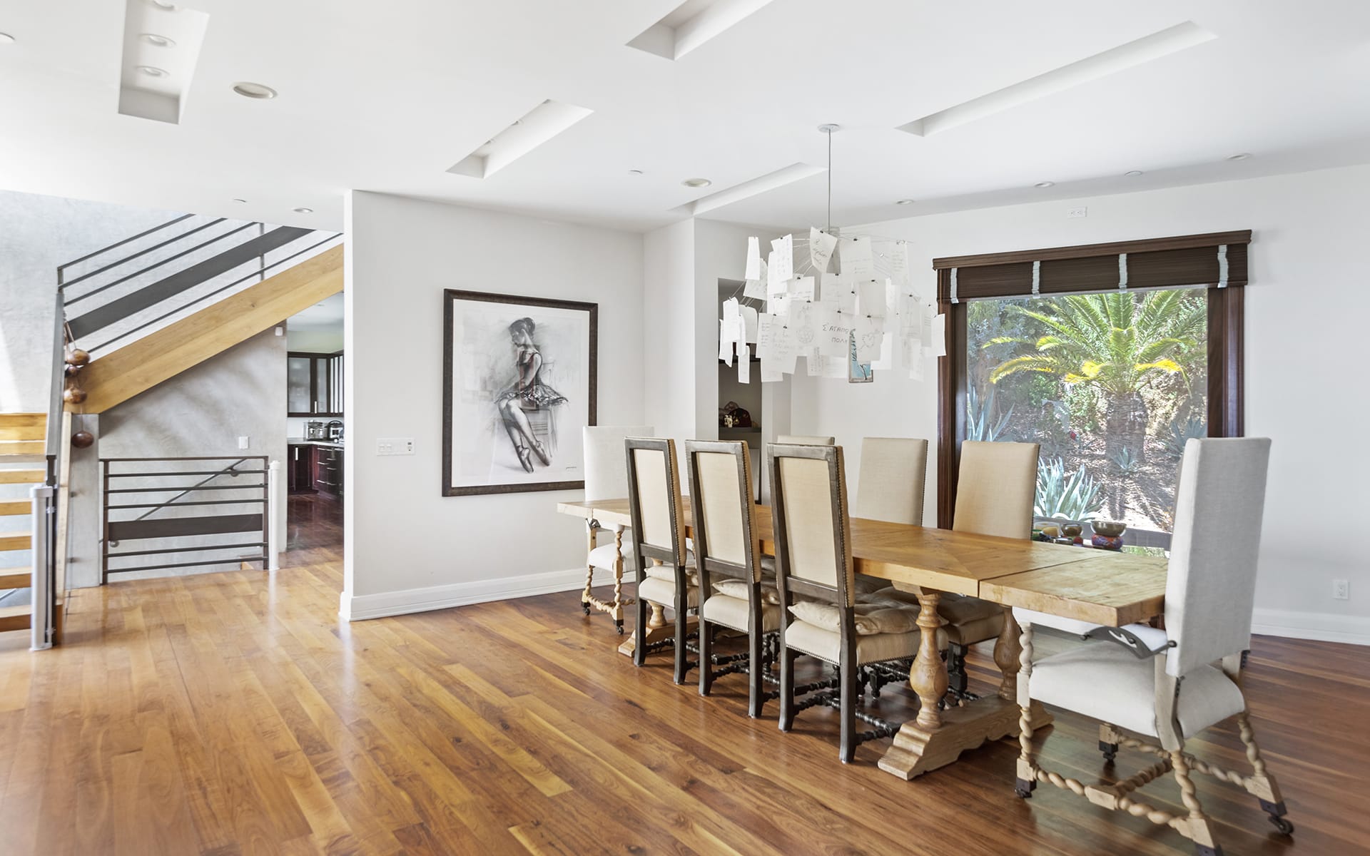 Modern dining room with a long wooden table, eight chairs, art on the wall, wooden floor, and unique ceiling light fixture.