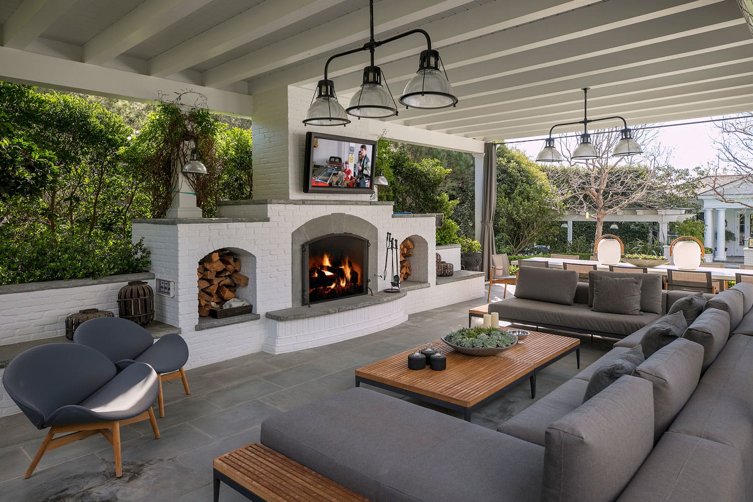 Outdoor patio with gray seating, wooden tables, and a white brick fireplace under a pergola, surrounded by greenery.