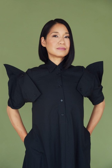 Person in a black dress with unique shoulder design stands confidently against a green background.