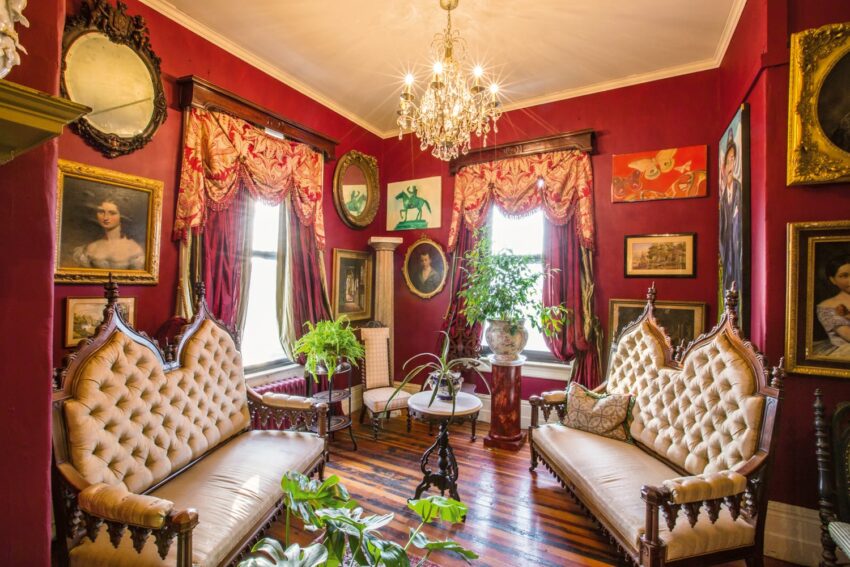 Victorian-style living room with chandeliers, ornate furniture, red walls, paintings, and plants.