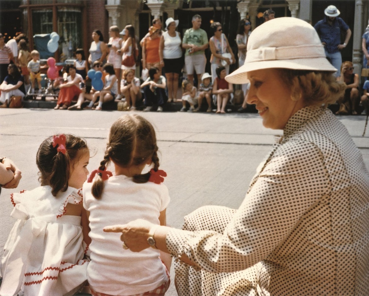 Woman in a hat sitting with two children at a parade, crowd and balloons in the background.