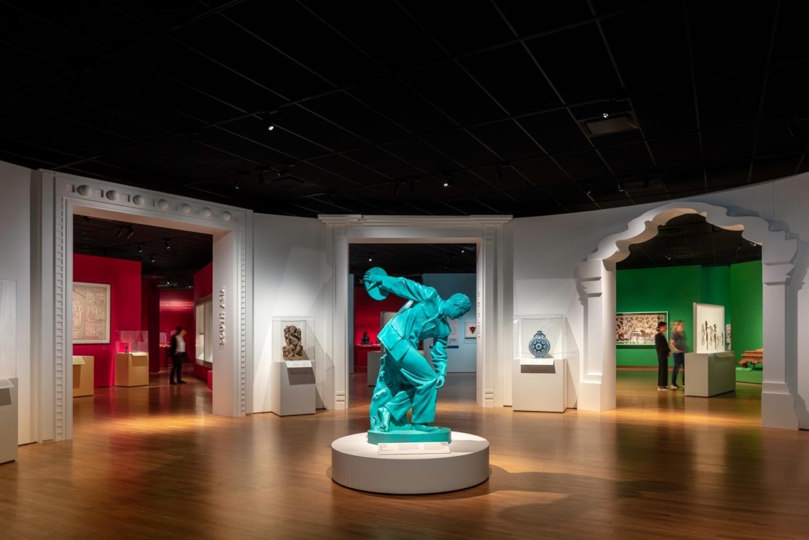 Exhibit hall with vibrant walls, various artworks, and a central turquoise statue on a pedestal.