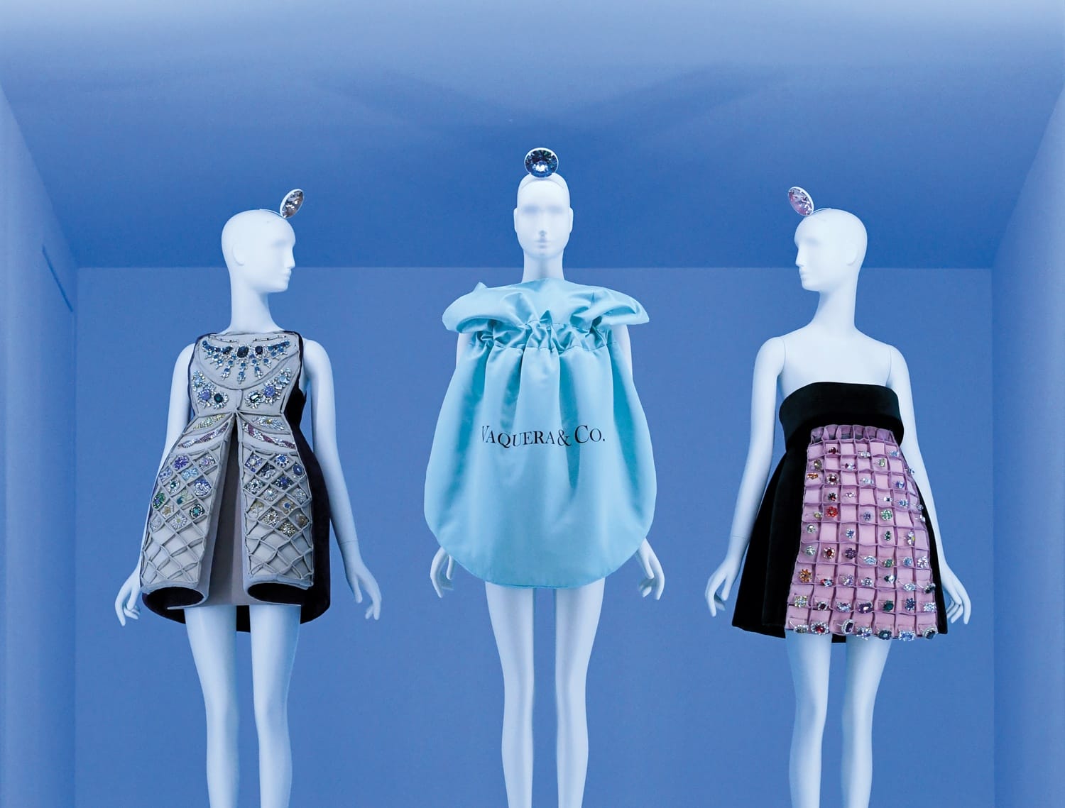 Three mannequins displaying fashion-forward dresses with unique design elements in a minimalist blue display room.