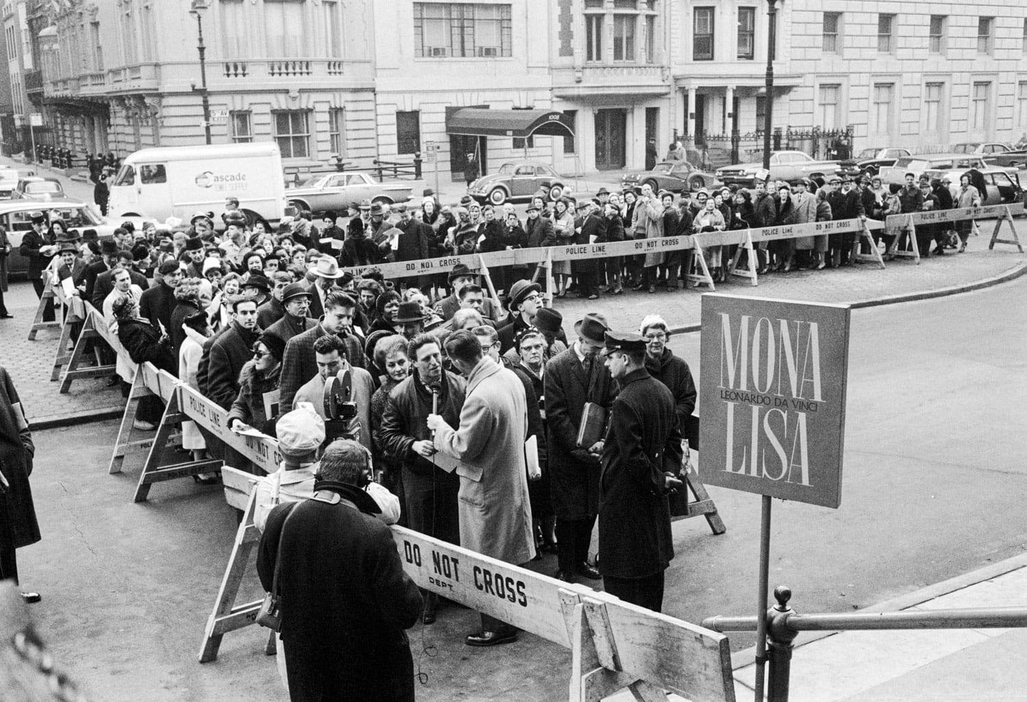Crowd of people in line outside a building for the Mona Lisa exhibition with barricades and a sign in an urban setting.