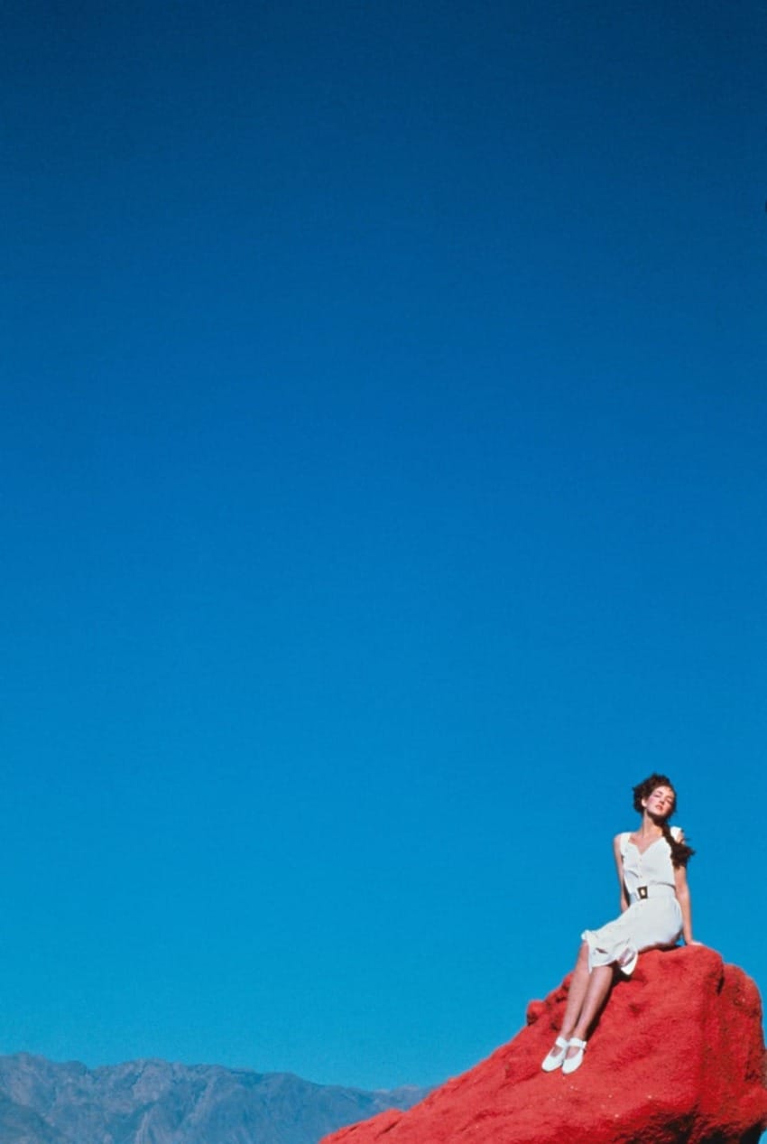 Woman sitting on a red rock against a clear blue sky in a mountainous landscape.