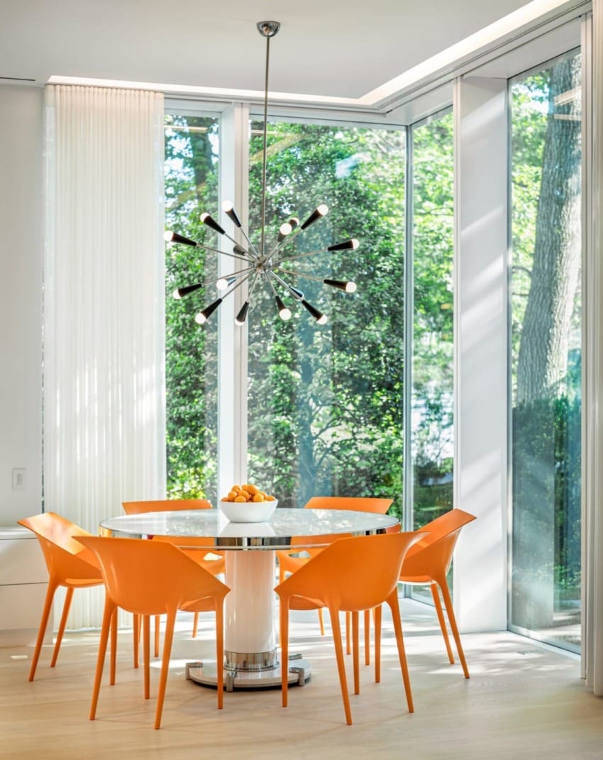 Modern dining room with orange chairs around a white table, large windows, sputnik chandelier, and forest view outside.