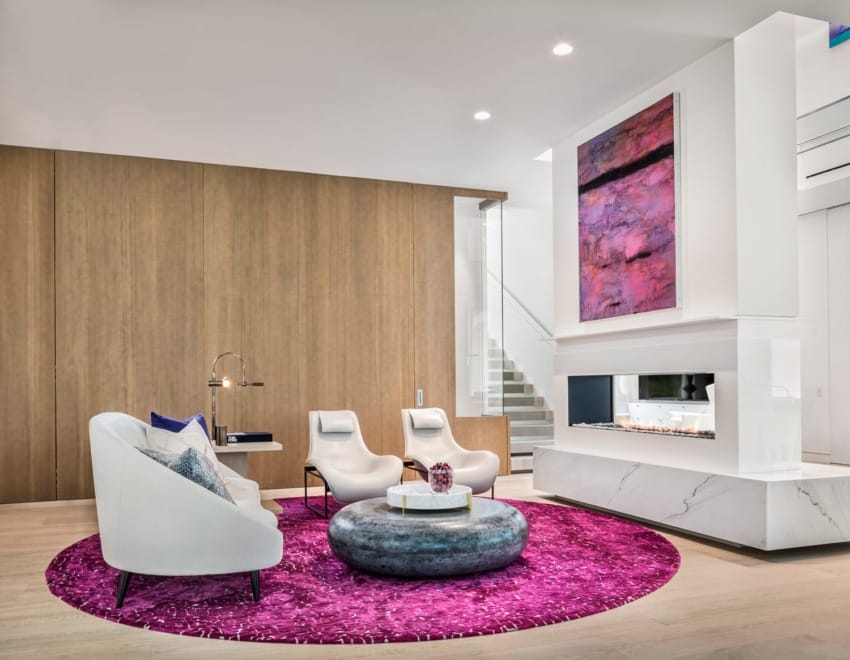 Modern living room with white chairs, round coffee table, pink rug, abstract art, and a contemporary fireplace.