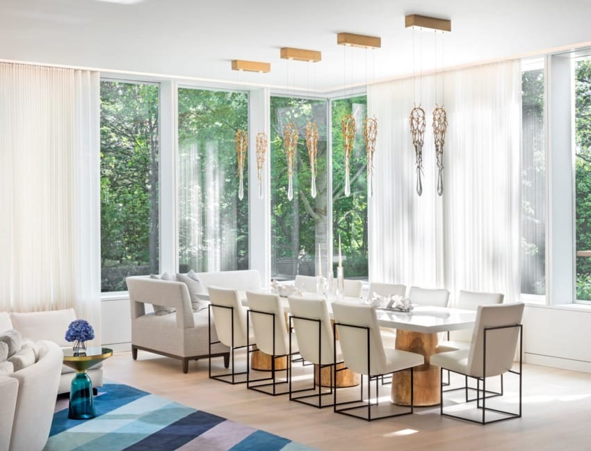 Modern dining room with large windows, white chairs, rectangular table, unique hanging lights, and colorful geometric rug.
