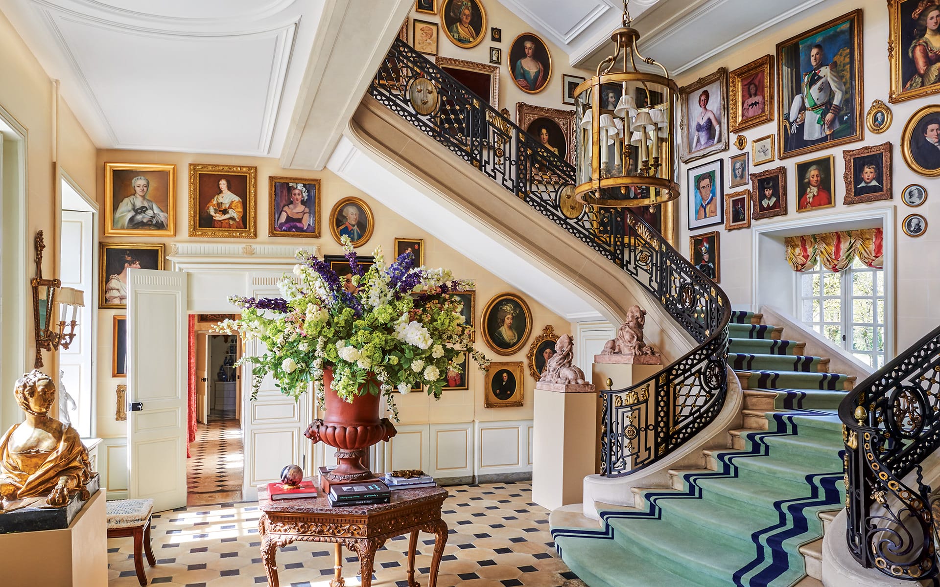 Elegant grand staircase with artwork-covered walls, floral arrangement, and checkered floor in a stately, sunlit interior.